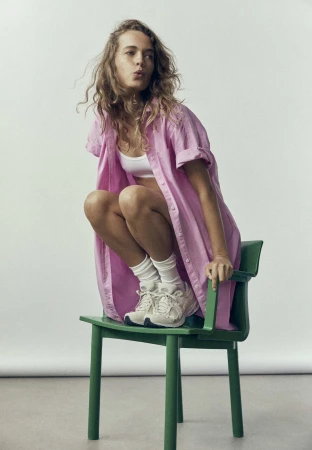 Woman in pink linen shirt crouching on a green chair wearing white sneakers against a white background