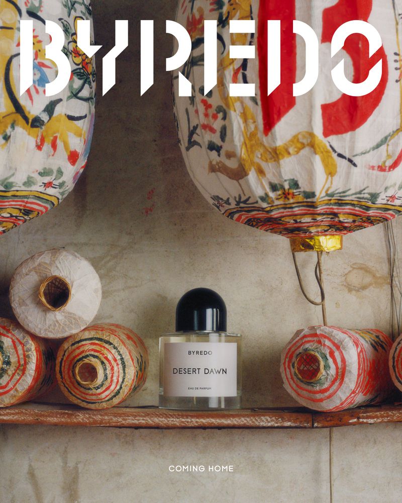 Byredo Desert Dawn perfume bottle centered on shelf, surrounded by patterned paper lanterns, against textured wall, warm color palette.