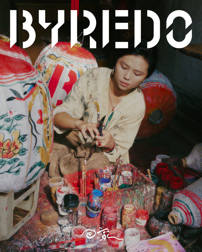 Asian woman artist painting designs on traditional lanterns in a workshop with paint jars and the brand BYREDO visible.