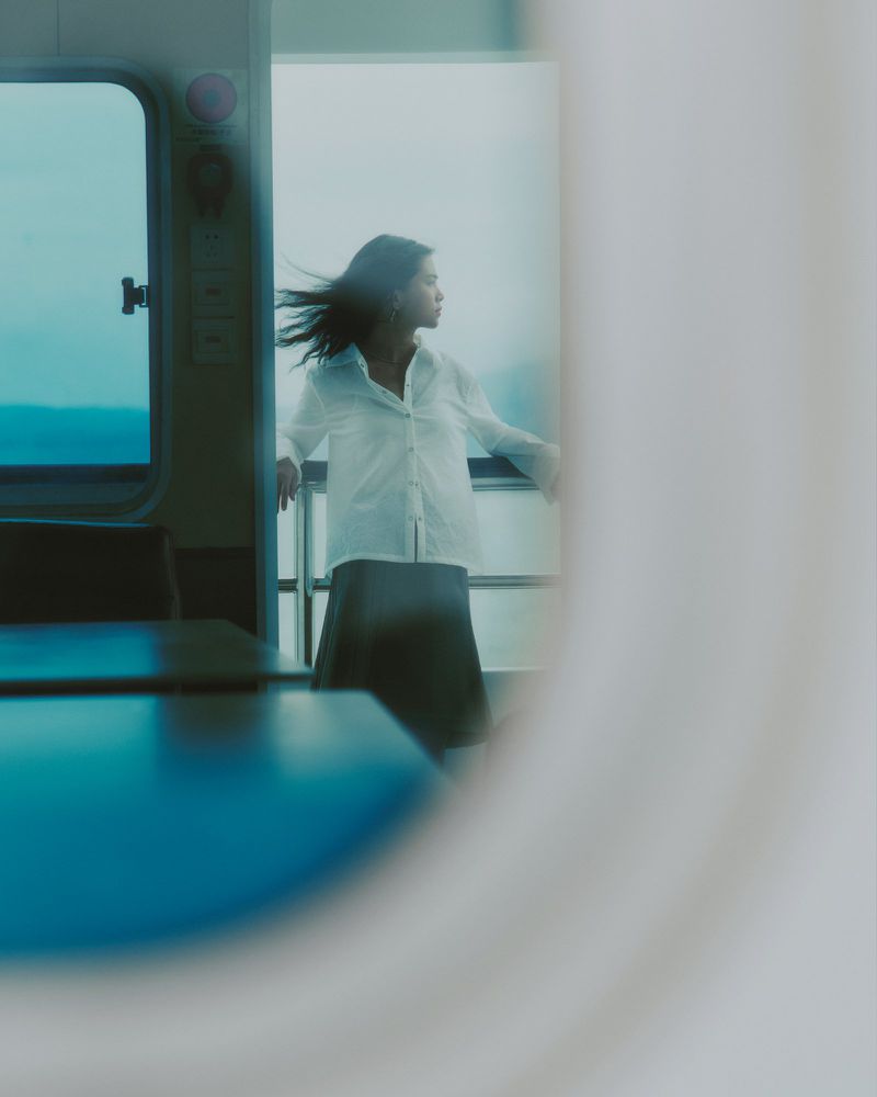 Adult woman with windblown hair wearing skirt and button-down shirt stands on deck with water view.