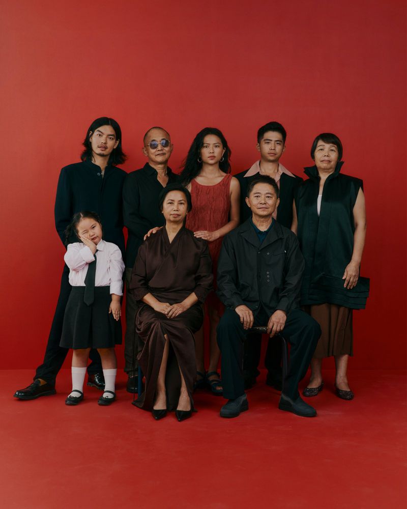 Group portrait of eight people against a vibrant red backdrop, featuring diverse ages and elegant attire, suitable for fashion or family themes.