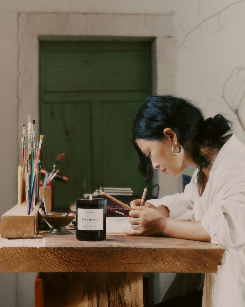 Woman with dark hair sits at a wooden desk, drawing with pencils, with art supplies, a lit candle, and a green door in background.