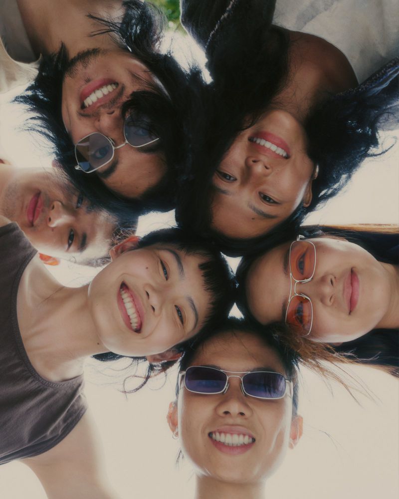 Low angle group portrait of six smiling young adults leaning in together in a tight circle against an open sky
