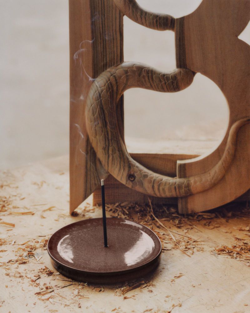 Studio shot of brown incense holder with incense stick and a light brown wood carving against a light neutral background.