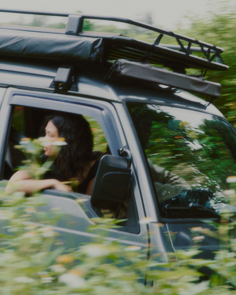 Woman driving a SUV with a roof rack through a green, blurred landscape. The vehicle is grey and surrounded by lush foliage.
