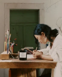 East Asian woman drawing at a wooden table next to a burning Byredo candle in an artist studio
