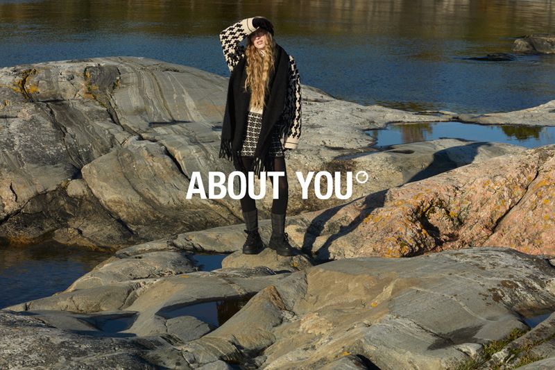 Woman in patterned hat and coat stands on rocky shore near water, posing for fashion shoot in outdoor setting during daylight hours.