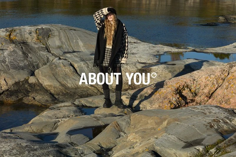 Woman in patterned hat and coat stands on rocky shore near water, posing for fashion shoot in outdoor setting during daylight hours.