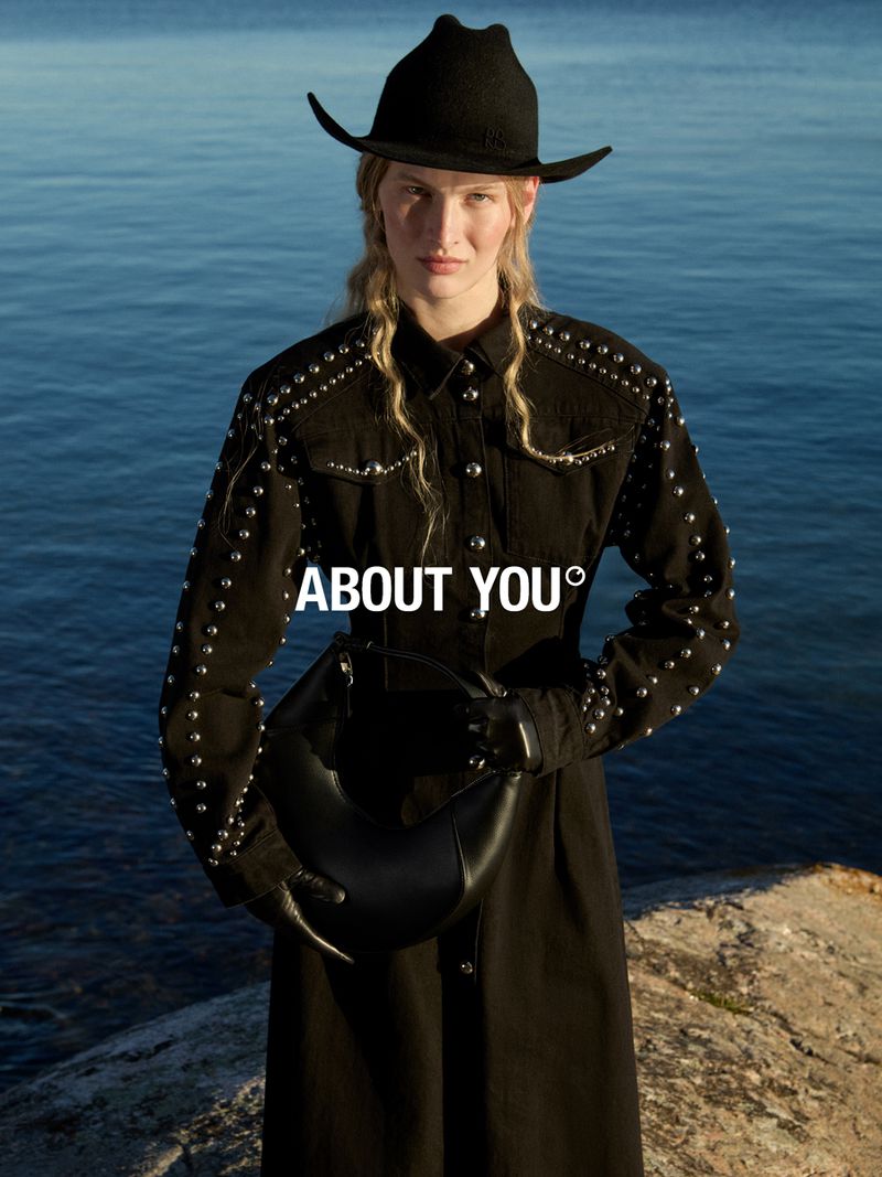 Woman poses with a black handbag against an ocean backdrop, wearing a studded black jacket and hat, in a fashion advertisement.