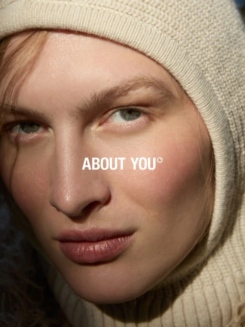 Close-up portrait of an adult female model with fair skin, wearing a hooded, knitted top, featuring the About You brand logo.