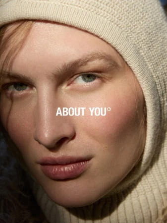 Close-up portrait of an adult female model with fair skin, wearing a hooded, knitted top, featuring the About You brand logo.