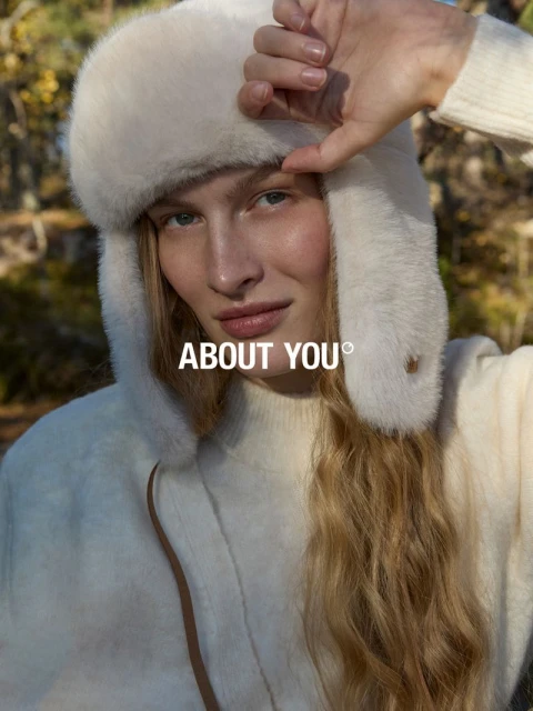 Fashion portrait of a blonde model in a white faux fur hat and jacket standing outside with the text “ABOUT YOU” overlaid.