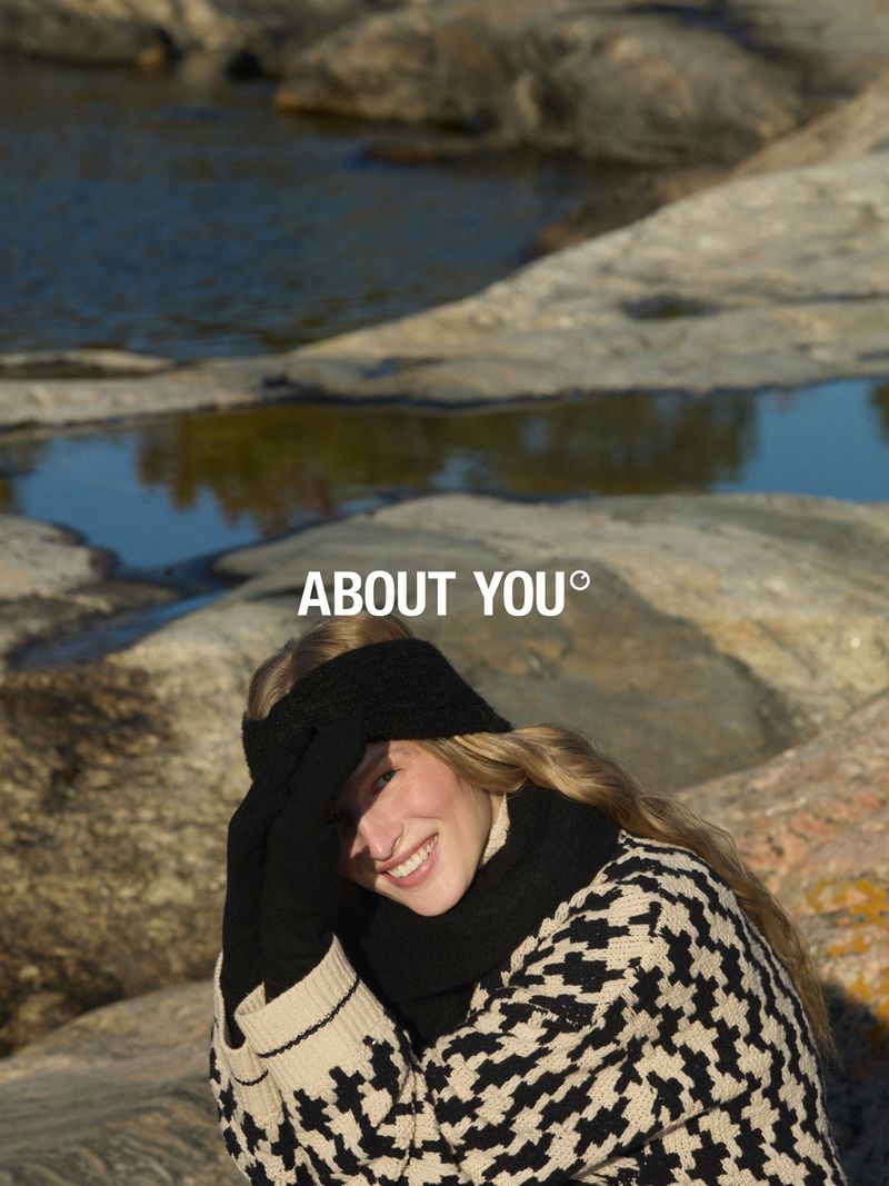 Smiling woman in black headband, gloves, and patterned sweater poses outdoors near rocks and water with the ABOUT YOU logo above.
