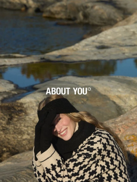 Smiling woman in black headband, gloves, and patterned sweater poses outdoors near rocks and water with the ABOUT YOU logo above.