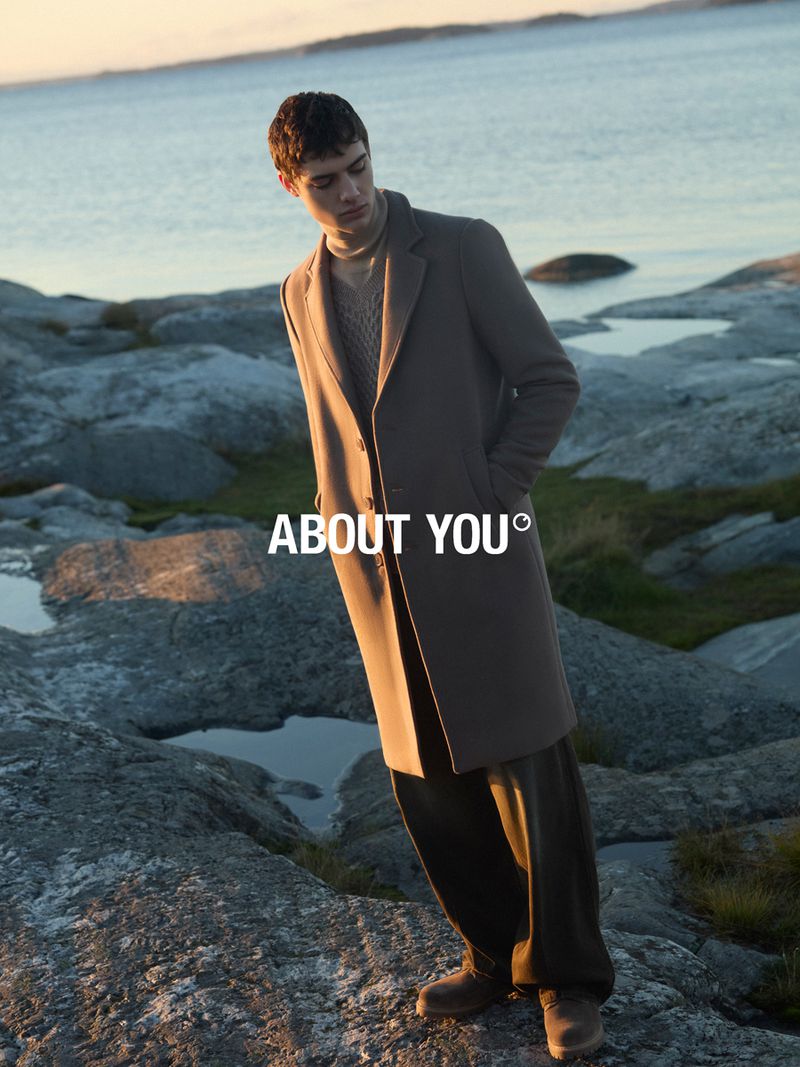 A young adult male model poses on a rocky shore in a brown coat and pants with the text ABOUT YOU overlaid in a fashion advertisement.