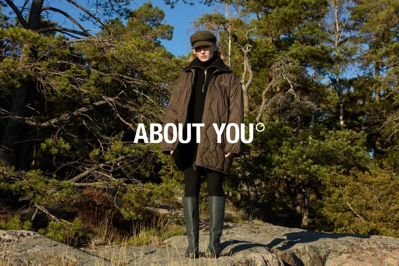 A woman in a quilted jacket, cap, and boots poses on rocks in a forest setting for an About You fashion advertisement.