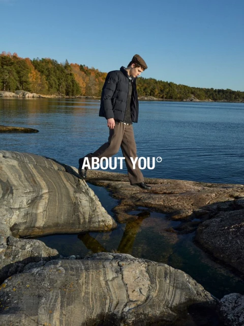 A man in a jacket and cap walks across rocks by the water on a sunny day, showcasing outdoor fashion with a nature background.