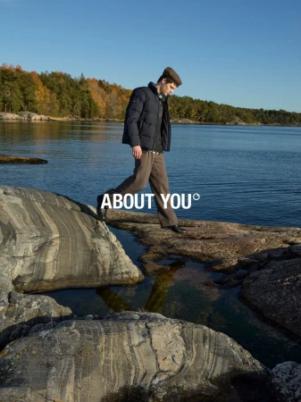 A man in a jacket and cap walks across rocks by the water on a sunny day, showcasing outdoor fashion with a nature background.
