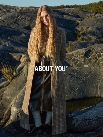 Model with long blonde hair wearing a plaid coat outdoors against a rocky coastal landscape background.