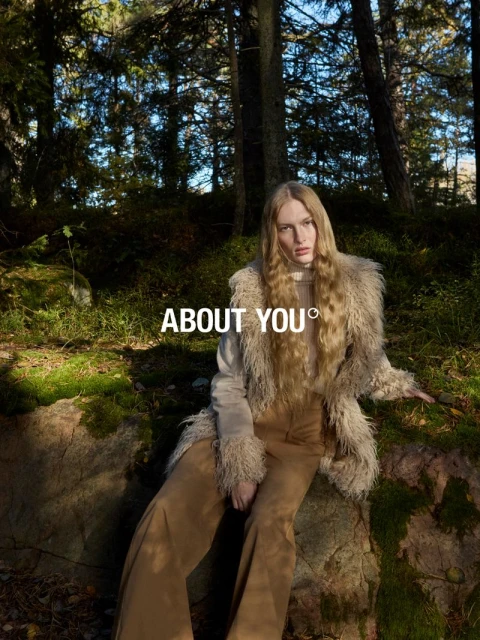 A fair-skinned model with long blonde hair poses while sitting on a rock in a forest, wearing brown pants and a fur-trimmed jacket.