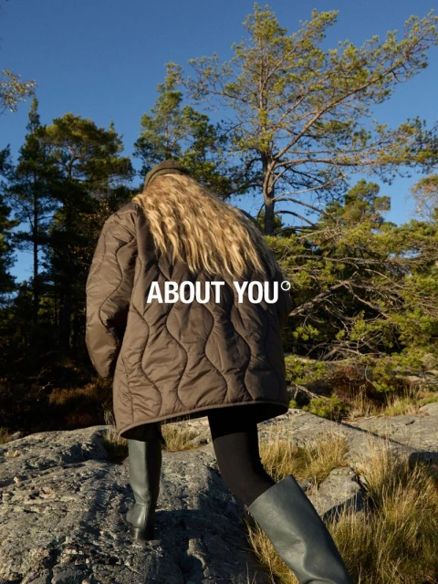 Woman in a brown quilted jacket and grey boots walks on a rocky path in a wooded area under a bright blue sky; fashion photography.