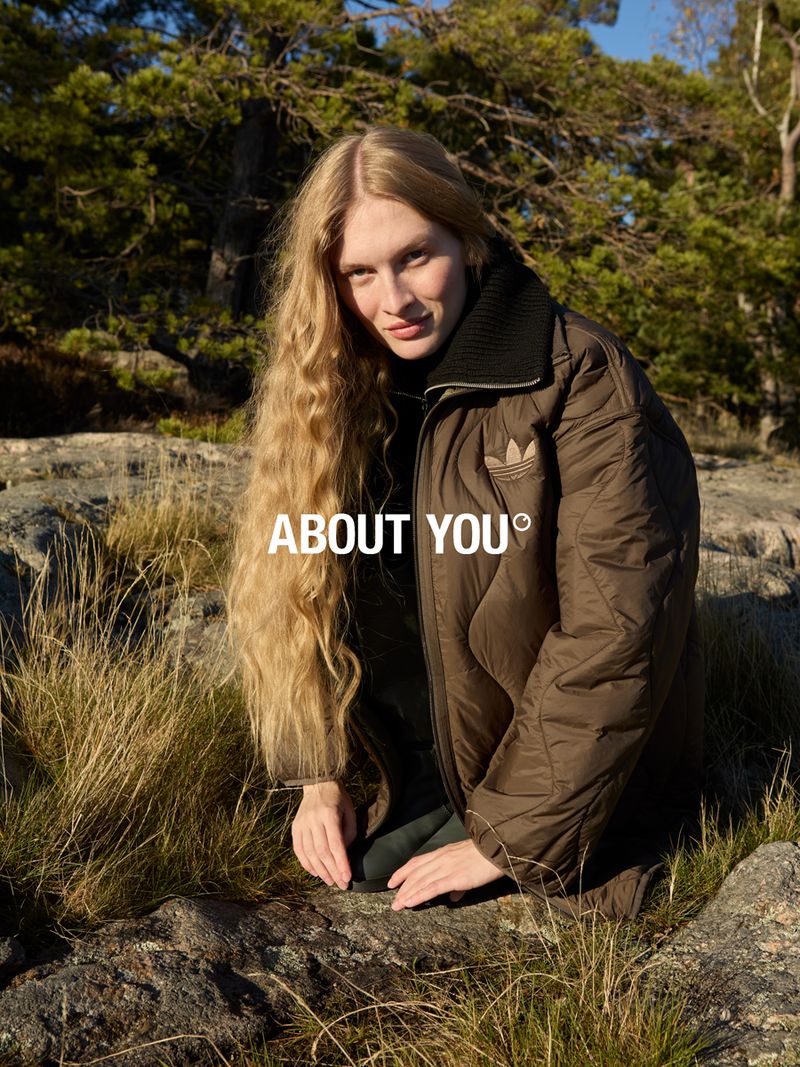 A blonde woman in a brown jacket poses on a rock in nature. The picture highlights earthy tones and an outdoor setting. "ABOUT YOU" text overlay.