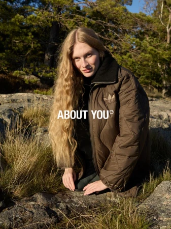 Model Astrid Baauer kneeling on rocky terrain wearing a brown puffer jacket for the About You FW25 campaign.