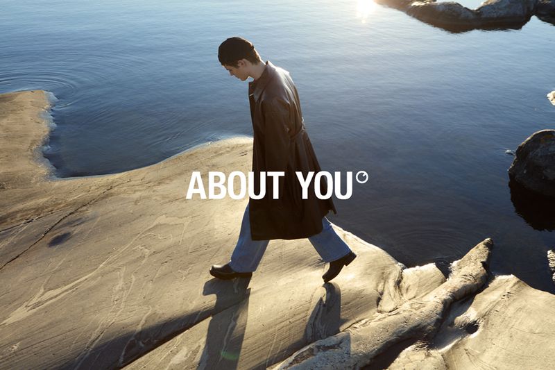 A fashionable person in a black coat and blue jeans walks on a rock formation near a body of water, with the 'About You' logo visible.