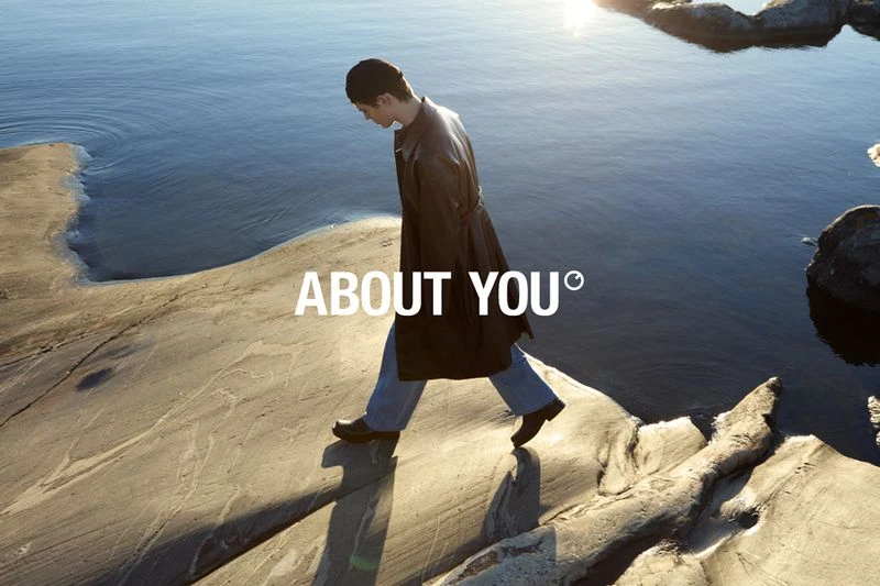 A fashionable person in a black coat and blue jeans walks on a rock formation near a body of water, with the 'About You' logo visible.