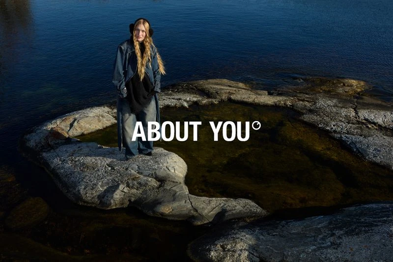 A young adult female stands on a rock formation in a river wearing a long denim jacket and wide-leg jeans with the ABOUT YOU logo.