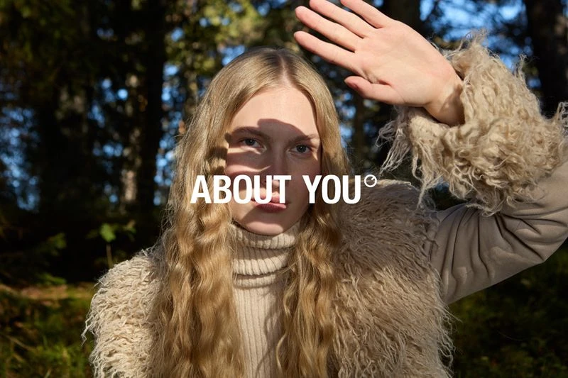 Portrait of blonde woman in a forest setting, shielding eyes with hand. She wears a beige turtleneck and fur-trimmed jacket.