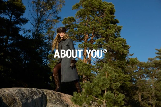 A woman in a coat and hat poses on a rock outcrop with trees and a blue sky in the background. Fashion and nature combine.