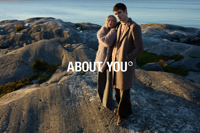 A stylish couple stands on a rocky shore near the ocean under a bright sky, promoting the brand 'About You' with a scenic backdrop.