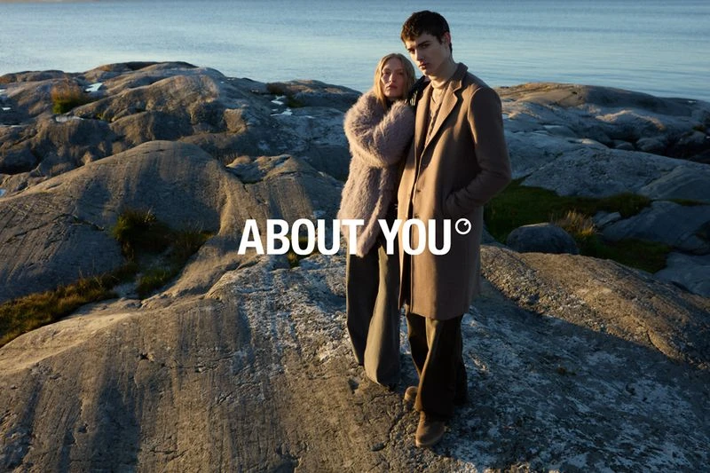 A stylish couple stands on a rocky shore near the ocean under a bright sky, promoting the brand 'About You' with a scenic backdrop.