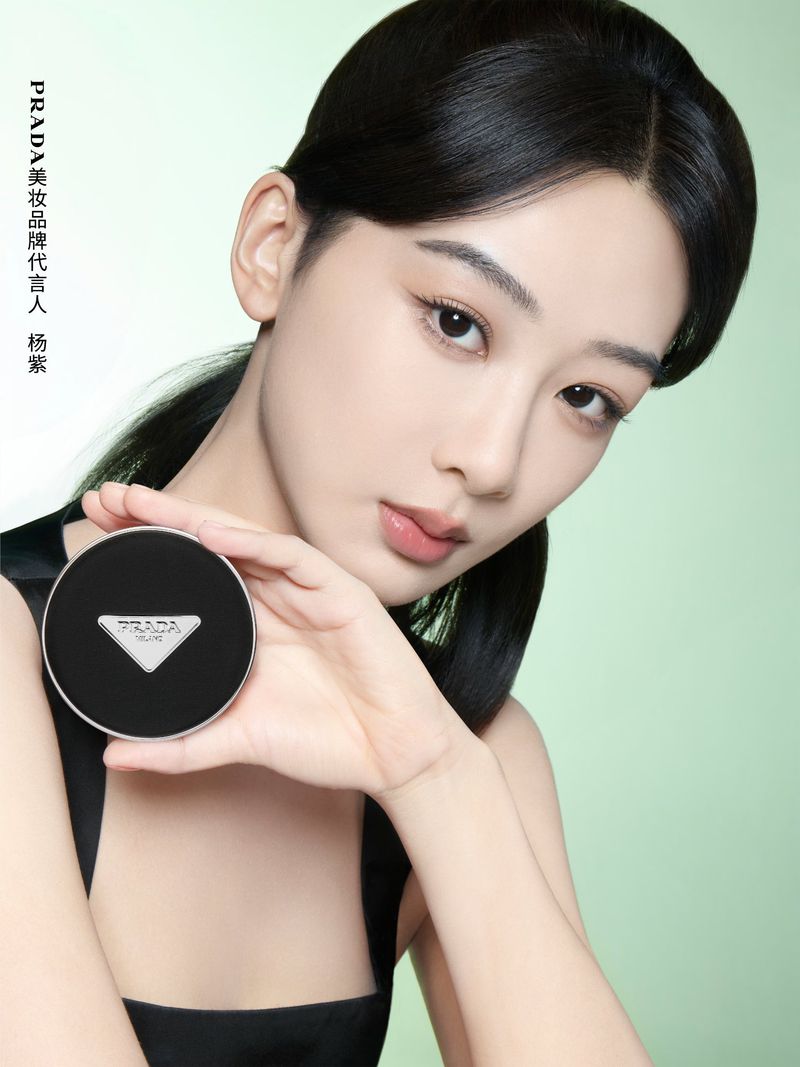 Close-up of a young Asian woman holding a Prada compact makeup case against a soft green backdrop in a beauty advertising campaign.