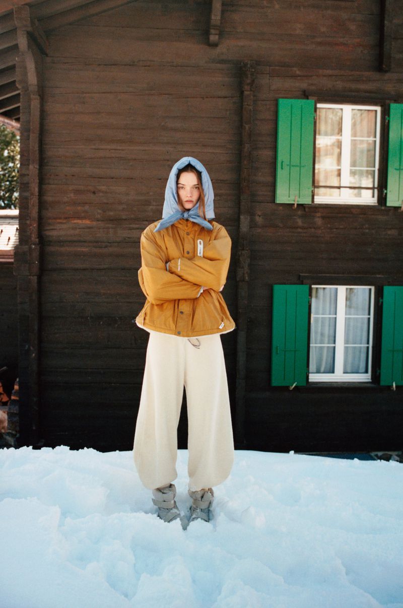 Woman in jacket and headscarf standing in snow in front of a wooden house with green shutters in wintertime.