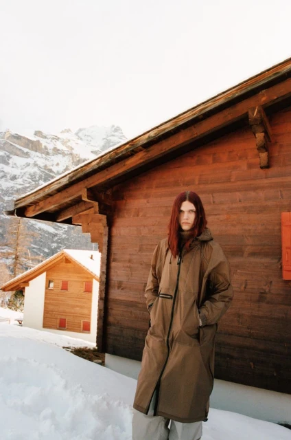 Adult female stands outside wooden cabin in snowy mountain landscape wearing a long, hooded brown coat and neutral pants in daylight.