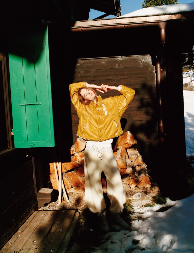 Woman in yellow jacket and cream pants poses near a wood cabin with stacked firewood in a snowy, sunlit environment during daytime.