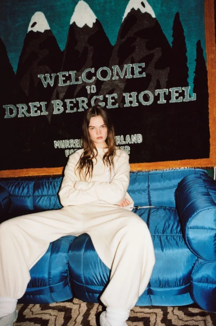 A young woman sits on a blue couch, wearing a beige sweatshirt and sweatpants with a mountain tapestry in the background.