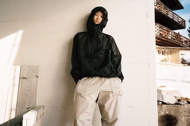 Asian man wearing a black hooded jacket and khaki pants stands against a white wall with a multi-story building in the background.