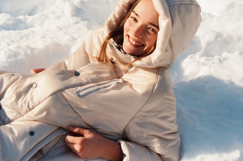 Smiling woman wearing hooded puffy jacket in a snowy winter landscape with soft, natural lighting and a shallow depth of field.