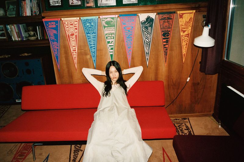 Young woman in a light-colored dress lounges on a red sofa in a wood-paneled room with pennants, casting a relaxed, casual mood.