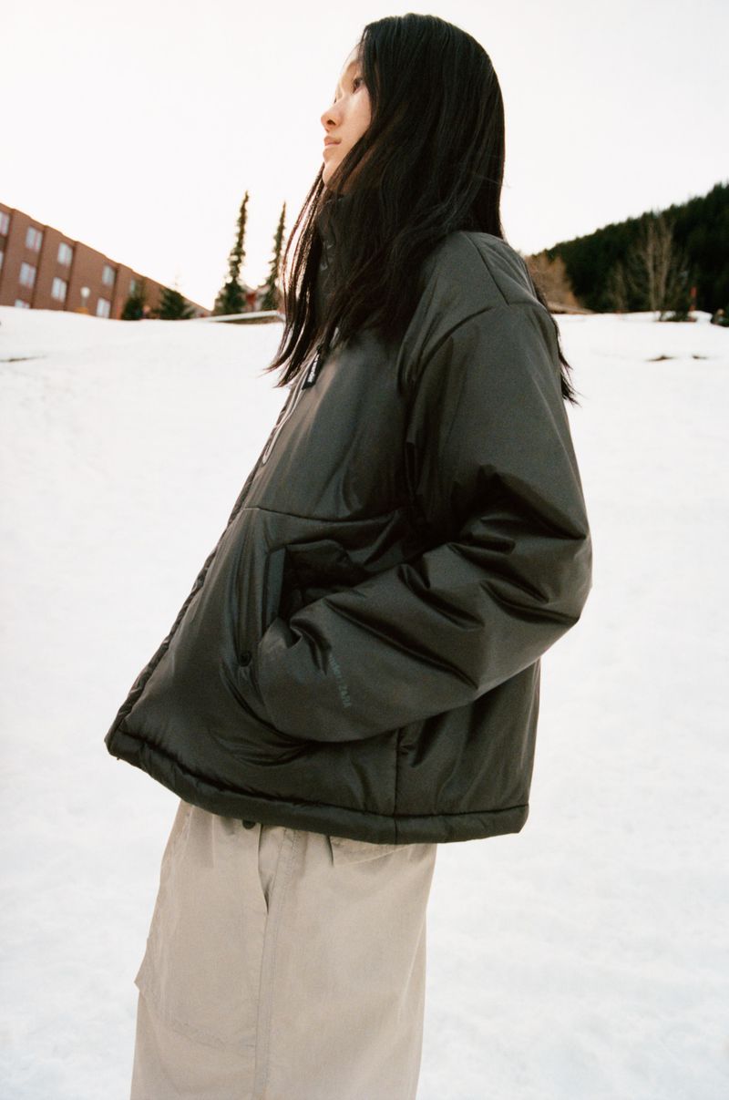 An Asian woman with long dark hair poses in snow wearing a black puffer jacket and beige pants, building and trees visible in background.