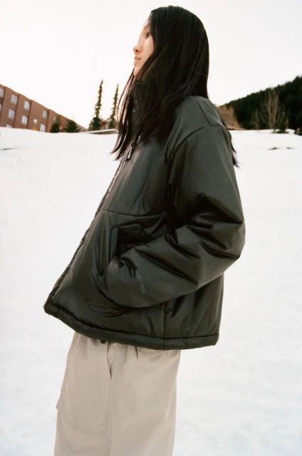 An Asian woman with long dark hair poses in snow wearing a black puffer jacket and beige pants, building and trees visible in background.