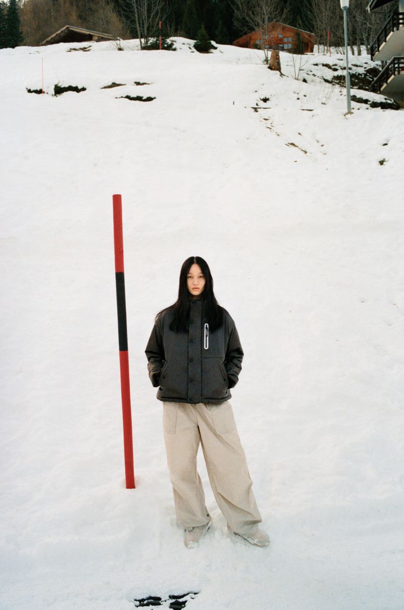 A full shot shows a slender young woman in winter apparel standing in snow, beside a red and black pole against a snowy hill backdrop.