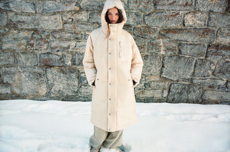 Model wearing a cream puffer coat with a hood standing against a rugged stone wall and snow-covered ground