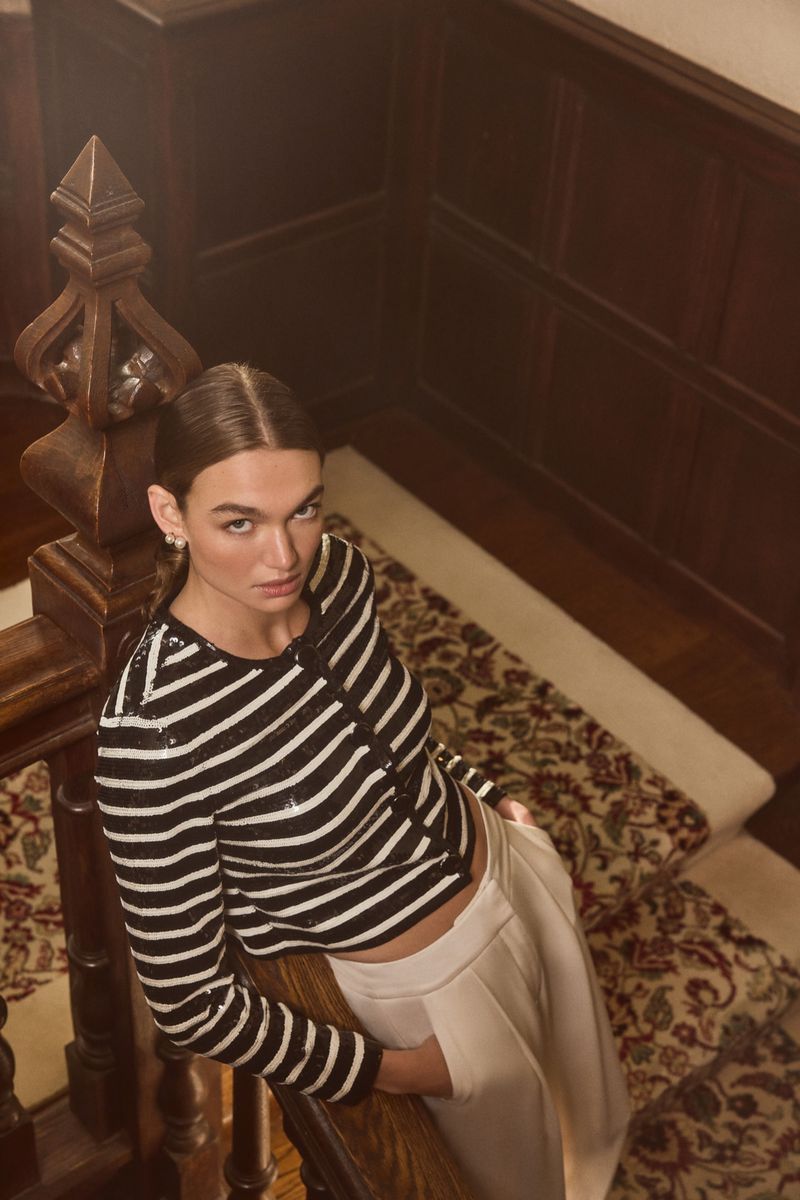 Adult woman with brown hair poses on a wooden staircase wearing a black and white striped long sleeve and white slacks.
