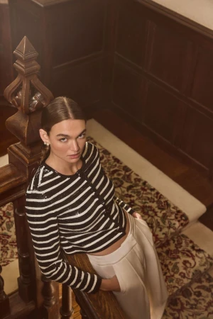 Adult woman with brown hair poses on a wooden staircase wearing a black and white striped long sleeve and white slacks.