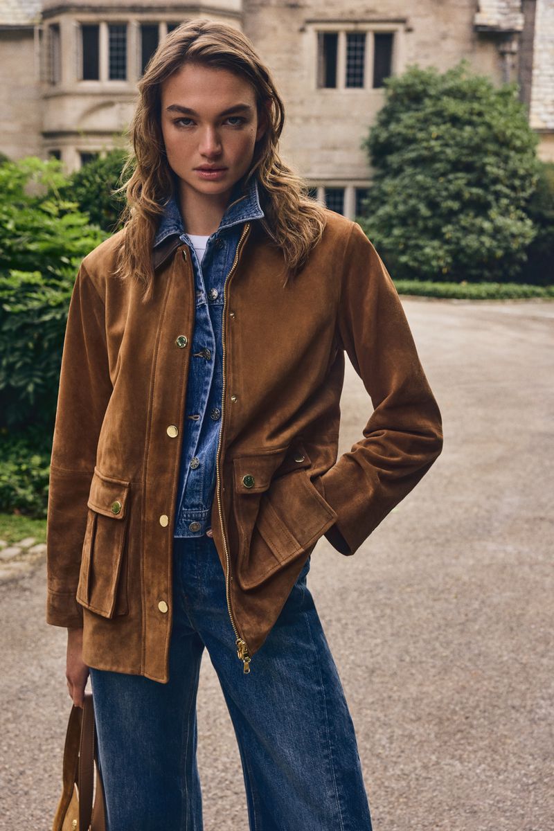 Model wearing a brown suede jacket over a denim jacket and jeans outdoors in front of a stone building.