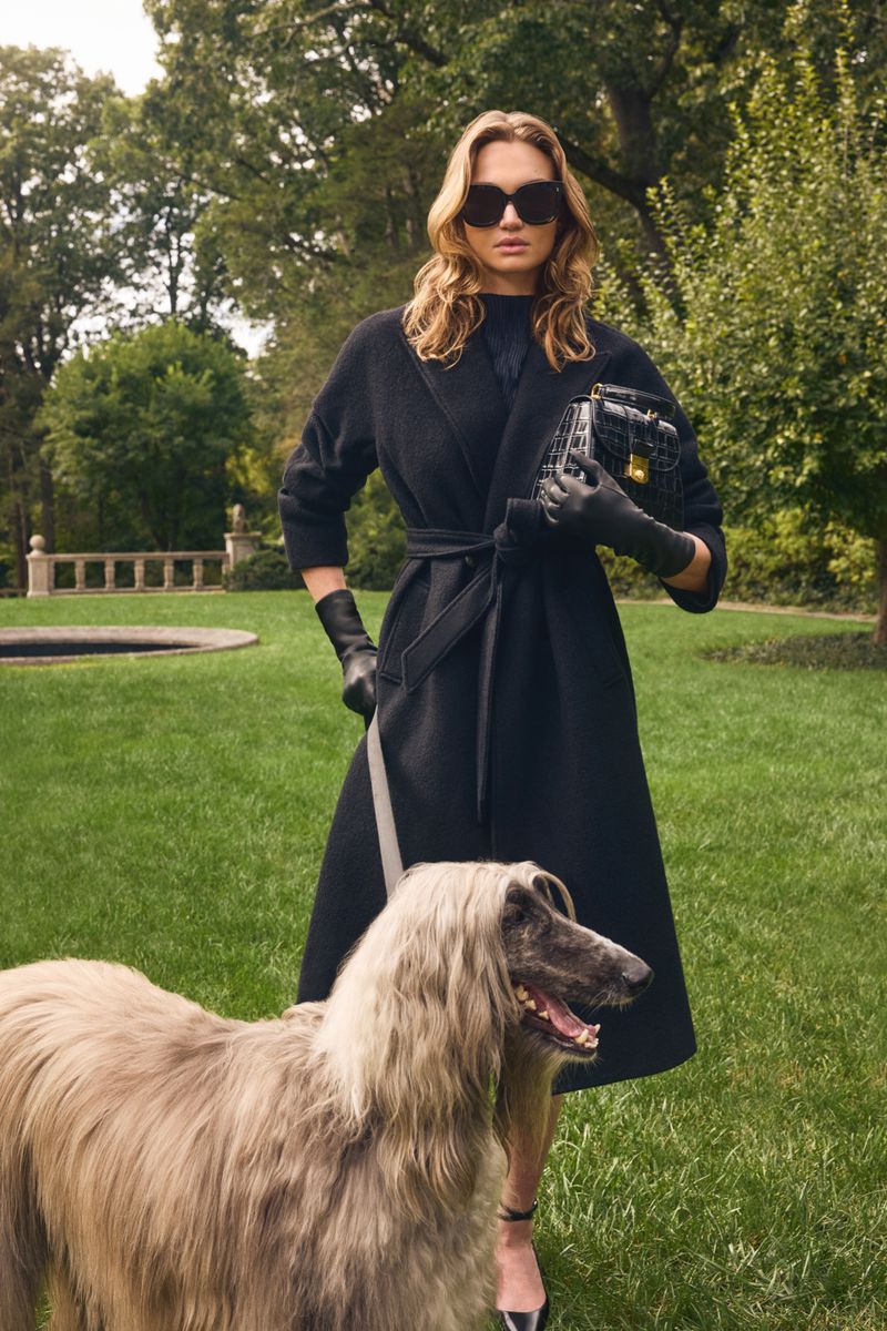 Woman in black coat walks an Afghan Hound on a leash in a grassy park setting with trees and a decorative stone feature.
